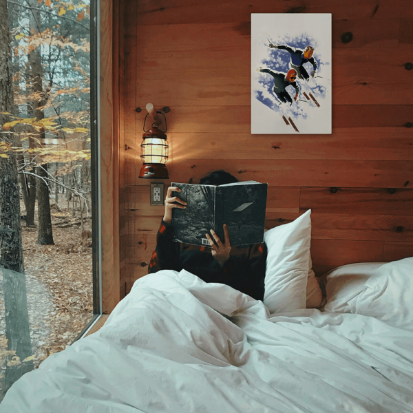 A Jantzen Vintage Ski poster with an image of two skiers on a white background hang above a woman in bed reading a book,