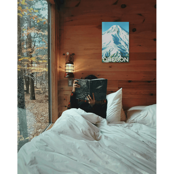 An aluminum sign print of Mt Hood hung on a cabin wall above a person in a bed reading.